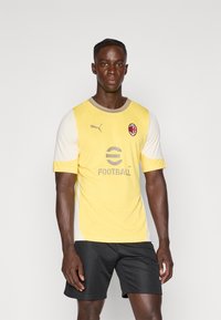 Maglia da calcio gialla e avorio con maniche corte, logo Puma, testo "eFootball" in grigio e emblema dell'AC Milan. Indossata con pantaloni corti neri.