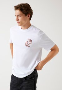 Camiseta blanca con mangas cortas, que presenta un logo gráfico en rojo y negro en el lado izquierdo del pecho. Hecha de algodón con una textura suave.