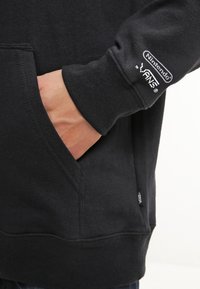 Sweat à capuche noir avec une poche avant, des poignets côtelés et un logo Nintendo sur la manche. Tissu doux avec une texture lisse.