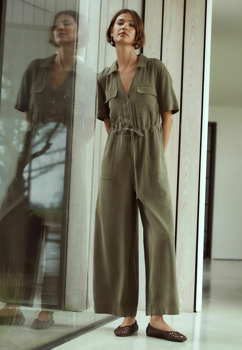 Femme aux cheveux courts portant une combinaison boutonnée vert olive et des ballerines tressées marron, debout à l'intérieur près d'une paroi vitrée reflétant son image.