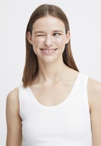 Ein weißes geripptes Tanktop hebt ein einfaches Design hervor, das einen runden Halsausschnitt und schmale Träger hat, getragen von einem Modell, das lächelt und dabei zwinkert.
