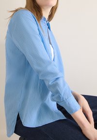 Femme portant une chemise boutonnée à fines rayures bleues et blanches par-dessus un haut blanc, assise avec les mains posées sur un jean foncé.