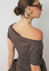 Abito marrone off-shoulder realizzato in tessuto morbido, con dettagli arricciati di lato e maniche corte. Accompagnato da orecchini audaci e occhiali da sole.