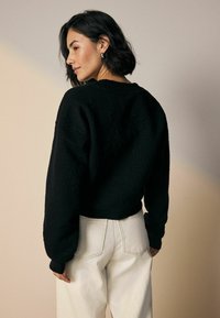 Frau mit kurzem dunklem Haar, die einen strukturierten schwarzen Pullover und weiße hochgeschnittene Jeans trägt und seitlich vor einem neutralen Hintergrund schaut.