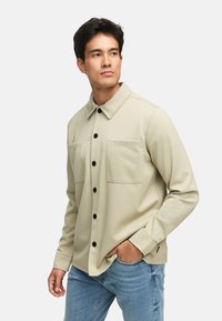 Lys beige knapp-up skjorte med krage, to frontlommer og svarte knapper; laget av teksturert stoff, kombinert med blå jeans.