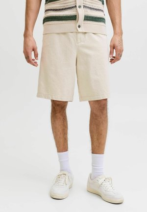 Mężczyzna w beżowych szortach, białych skarpetkach i białych trampkach, stojący z rękami swobodnie opuszczonymi wzdłuż ciała na jednolitym tle.