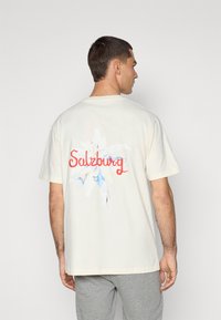 Krem majica s kratkim rukavima koja ima šareni cvjetni uzorak i natpis "Salzburg" na leđima. Izrađena od mekog materijala.