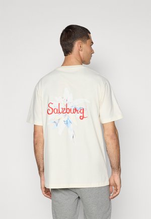 Krem majica s kratkim rukavima koja ima šareni cvjetni uzorak i natpis "Salzburg" na leđima. Izrađena od mekog materijala.