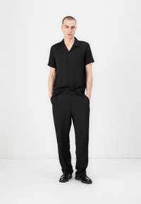 Chemise noire à manches courtes avec boutons et poche, assortie à un pantalon noir. Texture lisse, coupe décontractée, design minimaliste.