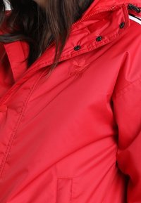 Rød isoleret jakke med lynlås og trykknaplukning, justerbar hætte og sidelommer. Har diskret logo broderi og kontrasterende detaljer.