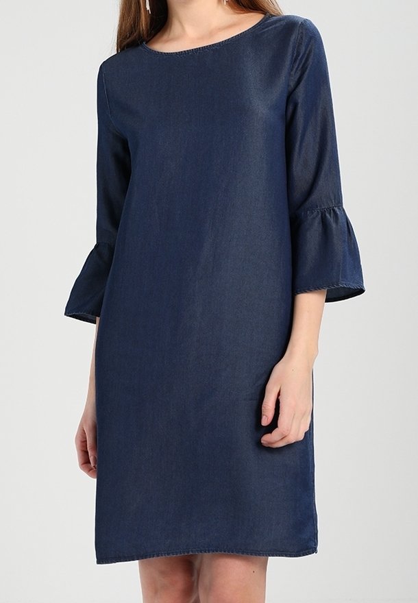 Femme portant une robe en denim bleu foncé longueur genou avec manches trois-quarts évasées et un col rond, sur un fond uni.