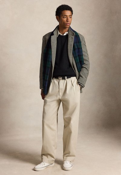 Hombre con pantalones beige, suéter negro, abrigo gris con espiga, bufanda de cuadros y zapatillas blancas de pie contra un fondo liso.