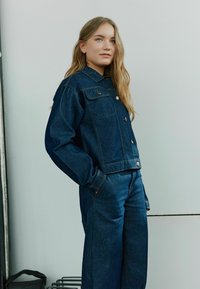 Denim jakna s ovratnikom, dvema žepoma na prsih in gumbom za zapiranje. Ima sproščen kroj in temno modro barvo, usklajena z ustreznimi jeans hlačami.