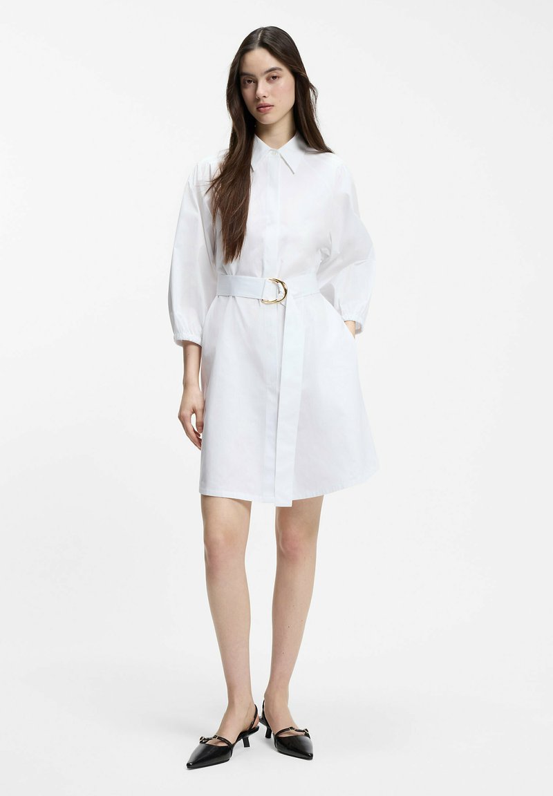 Femme debout portant une robe chemise blanche avec des manches bouffantes, une taille ceinturée et des chaussures noires à talons bas et bouts pointus sur fond blanc.