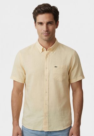 Uomo con capelli castani corti che indossa una camicia a maniche corte giallo pallido con bottoni e jeans azzurri, in piedi davanti a uno sfondo semplice.