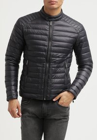 Veste matelassée noire avec fermeture éclair sur le devant, design matelassé, col montant et deux poches latérales zippées. Finition lisse et brillante avec poignets côtelés.