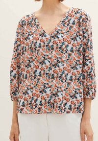 Blouse florale avec un col en V, ornée d'un motif de fleurs orange, turquoise et bleu foncé sur un fond blanc ; manches trois-quarts.