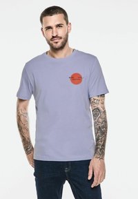 T-shirt en coton violet clair avec un accent rouge rond et un texte noir. Manches courtes, design encolure ras du cou, porté avec un jean en denim noir.