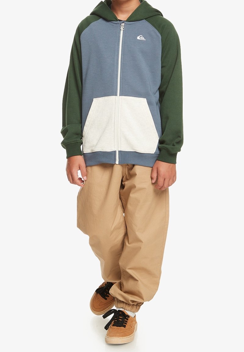 Jeune personne portant un sweat à capuche zippé bleu et vert avec une poche blanche, un pantalon ample beige, et des baskets marron clair avec des lacets noirs, debout sur un fond blanc.