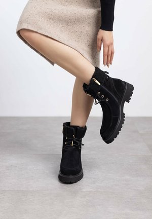 Botines de ante negro con suelas de goma gruesa, acentos de hardware dorado y un suave cuello de piel. Presenta cordones y detalles de cremallera lateral.