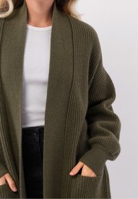 Oliwkowy kardigan z prążkowanej tkaniny, z otwartym przodem i długimi rękawami. Posiada boczne kieszenie oraz luźny krój. Noszony na białym topie.