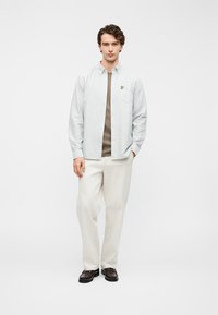 Μπλε ριγέ πουκάμισο με κουμπιά σε ανοιχτό μπλε και λευκό, συνδυασμένο με μπεζ μπλουζάκι, εκρού παντελόνι και σκούρα καφέ παπούτσια. Διακοσμητικό τσεπάκι στο στήθος.