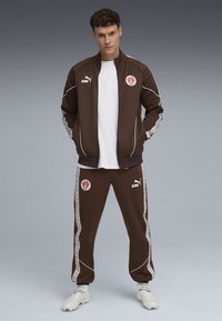 Brun atletisk træningsdragt med en zip-up jakke med hvide detaljer og logo, suppleret med matchende bukser, begge lavet af glat stof.