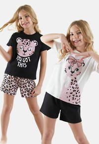 Dos chicas llevan atuendos a juego: una viste una camiseta negra con una cara de leopardo rosa y el texto "TÚ PUEDES"; la otra lleva una camiseta de leopardo blanca y pantalones cortos negros.