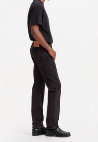Jeans de mezclilla negra con corte recto, que presentan un parche de cuero en la parte trasera, llevados con una camiseta negra de cuello redondo y botas negras.