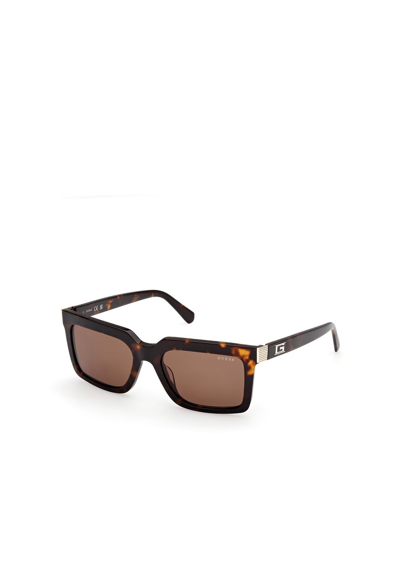 Occhiali da sole tartarugati con lenti marroni, montatura quadrata e dettagli metallici sulle aste. Presenta il testo "GUESS" all'interno della montatura.