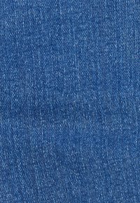 Tissu en denim, couleur bleu moyen avec un tissage en sergé, présentant une surface texturée et des variations subtiles de teinte et de fil.