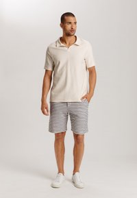 Camiseta polo corta de color crema con cuello, combinada con pantalones cortos a rayas en azul y blanco. Zapatillas blancas completan el conjunto.