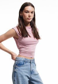 Jonge vrouw met lang bruin haar in een mouwloze roze top en hoog op de taille zittende blauwe jeans, staand met haar hand op haar heup tegen een witte achtergrond.