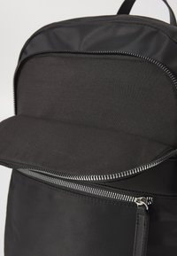 Black nylon backpack with a zippered front pocket and a smooth finish. The top flap is slightly open, revealing the interior lining.