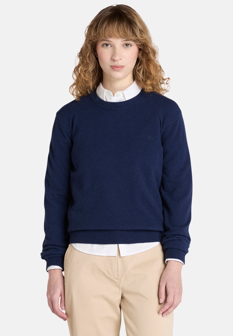 Granatowy sweter z okrągłym dekoltem wykonany z miękkiego materiału, z ribbingu na mankietach i u dołu. Noszony na białej koszuli z kołnierzykiem, o luźnym kroju.