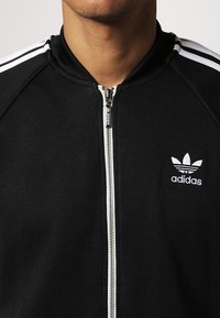Black zip-up jacket made from textured fabric, featuring white three-stripe accents on shoulders and a small white Adidas logo on the chest.