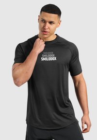Camiseta atlética negra de manga corta que presenta la palabra "SMILODOX" en diferentes tonalidades de blanco en la parte delantera. Textura suave, diseño ligeramente ajustado.