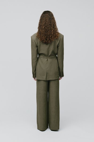 Personne aux longs cheveux bouclés, vue de dos, portant un costume ceinturé vert olive avec un pantalon à larges jambes, sur un fond uni.