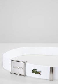 White elastic belt with a silver rectangular buckle featuring 'LACOSTE' logo; includes green embroidered crocodile detail on the strap.