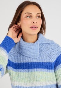 Maglione azzurro chiaro con colletto alto, caratterizzato da strisce orizzontali in tonalità di blu e verde. Maglia morbida e testurizzata con una vestibilità rilassata.