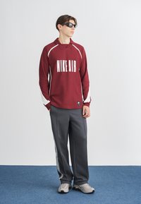 Camisa de manga longa bordô com o logótipo "NIKE AIR", detalhes brancos, combinada com calças de treino cinzas com riscas laterais e sapatos de desporto cinzentos.