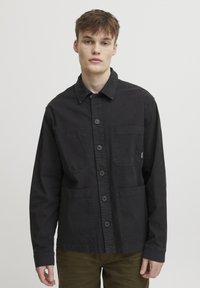 Black cotton jacket with a collared neckline, front button closure, two chest pockets, and long sleeves. Smooth texture, minimalist design.