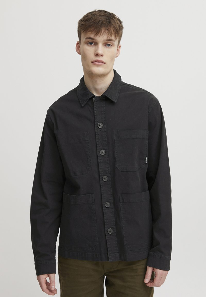 Black cotton jacket with a collared neckline, front button closure, two chest pockets, and long sleeves. Smooth texture, minimalist design.