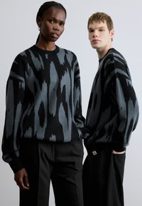 Dva manekena nosita prevelike puloverje z abstraktnim vzorcem v črni in sivi barvi. Puloverji imajo okrogle izreze in so izdelani iz teksturirane tkanine.