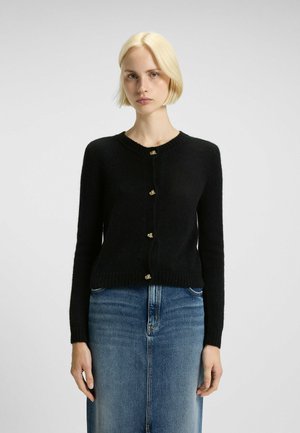 Black cardigan made of soft material, with a ribbed hem and neckline, featuring gold button accents down the center, paired with a denim skirt.