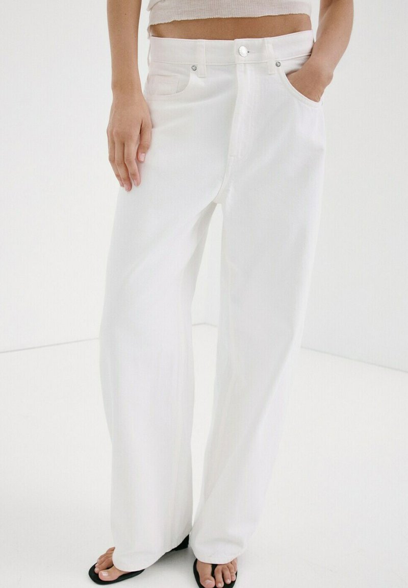 Personne portant un jean blanc taille haute à jambes larges, une main dans la poche, et des sandales noires à bouts ouverts, sur un fond blanc.