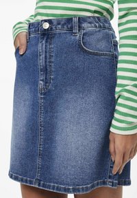 Lähikuva henkilöstä, joka käyttää sinistä farkkuhameita ja vihreä-valkoista raidoitettua pitkähihasta toppia, yksi käsi taskussa.