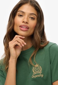 T-shirt en coton vert avec un col rond, des manches courtes et un logo brodé en or avec un motif de couronne et de laurier sur la poitrine.