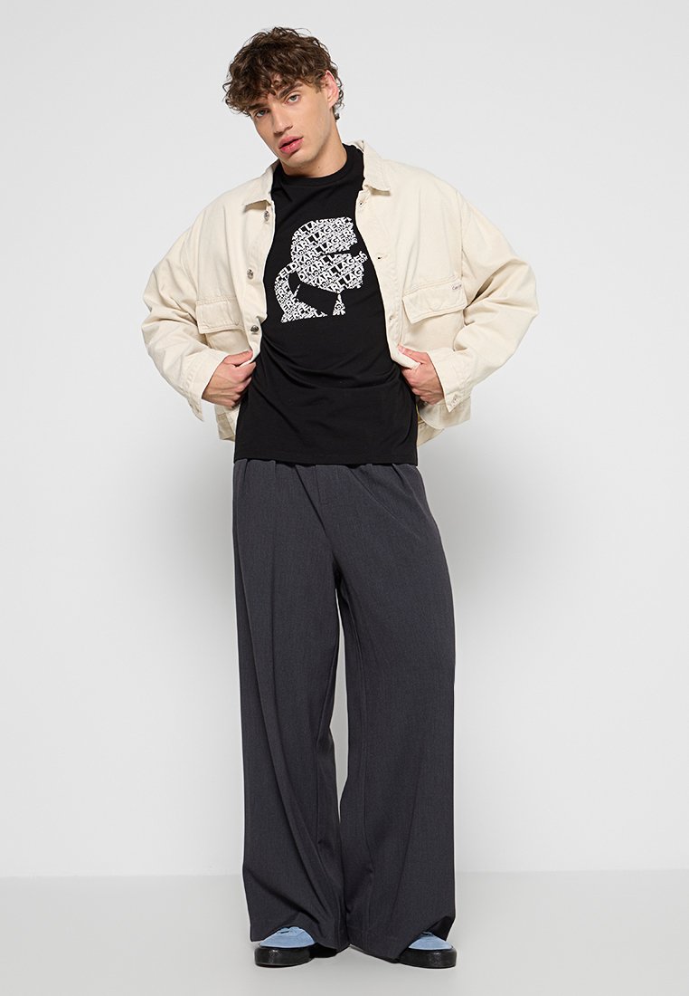 Chaqueta beige de gran tamaño, camiseta negra con estampado gráfico, pantalones anchos grises y zapatillas azules. Tejido suave con corte relajado.