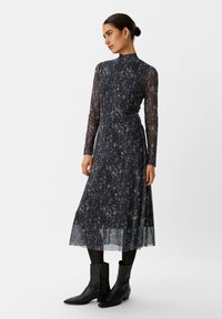 Vestito nero a maniche lunghe con un motivo testurizzato in diverse tonalità di bianco, caratterizzato da un collo alto e maniche trasparenti. Abbinato a stivali da cowboy neri.